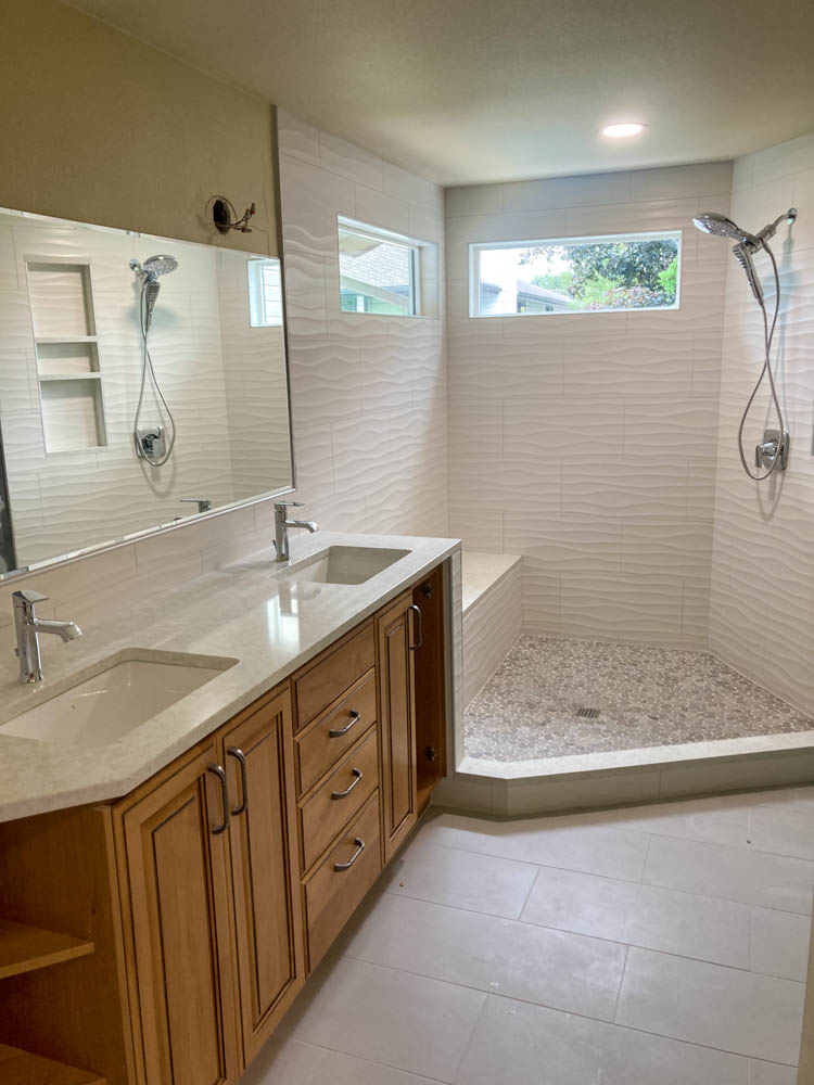 A modern bathroom with vanity cabinet with countertop and dual sink with faucet; a shower area with installed shower fixture and faucet
